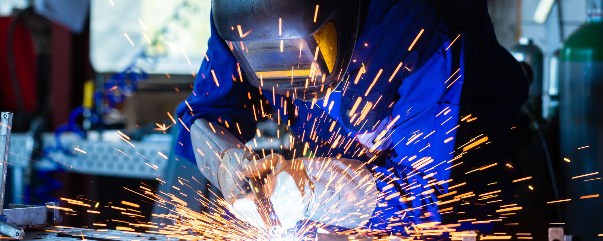 Welder bonding metal with welding device in workshop, lots of sparks to be seen, he wears welding goggles